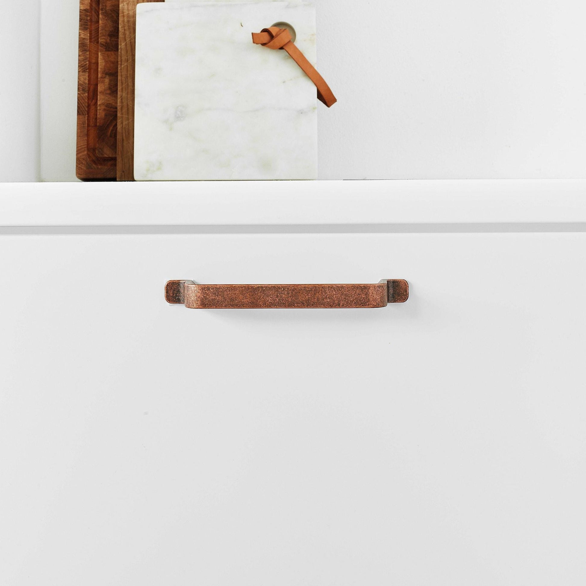 May include: A close-up of a copper cabinet pull with a slightly curved shape. The pull is mounted on a white cabinet door.