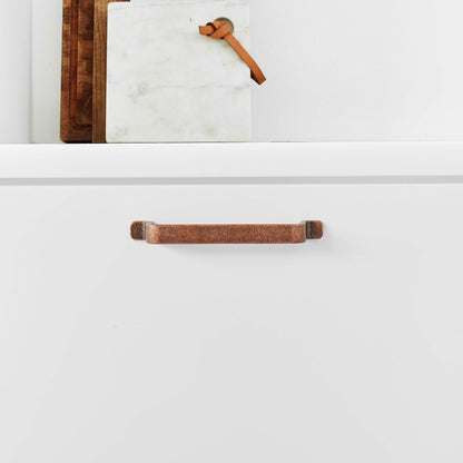 May include: A close-up of a copper cabinet pull with a slightly curved shape. The pull is mounted on a white cabinet door.