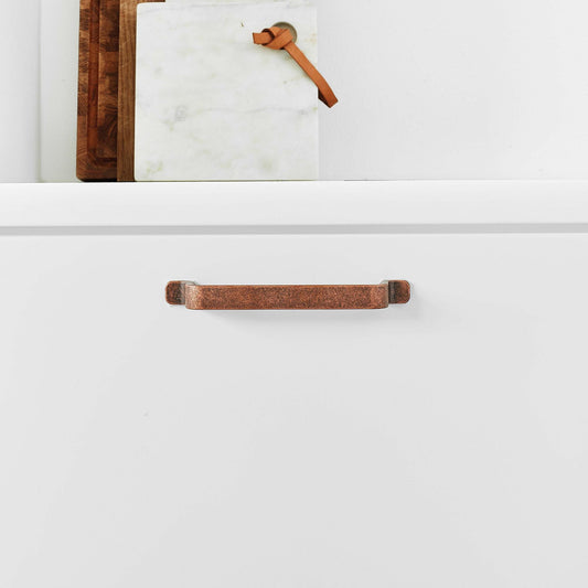 May include: A close-up of a copper cabinet pull with a slightly curved shape. The pull is mounted on a white cabinet door.